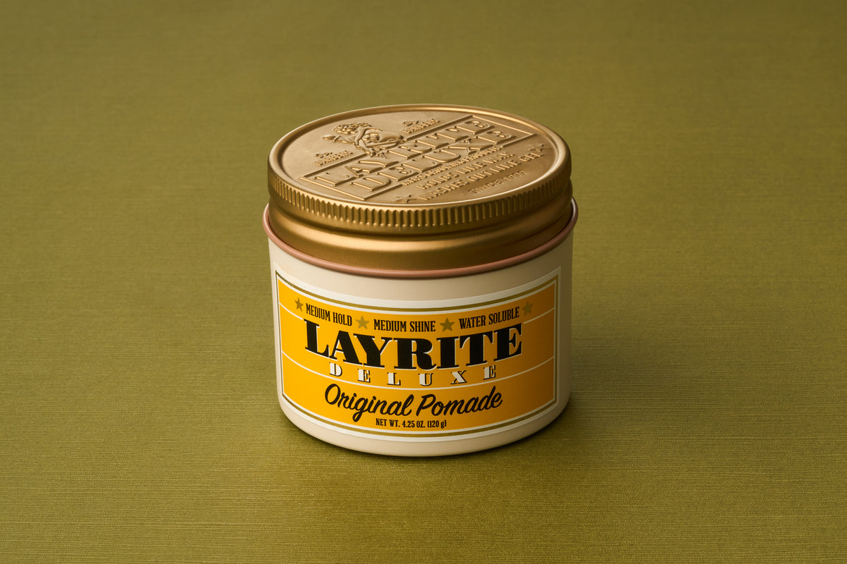 A jar of Layrite Original Pomade with a gold lid rests on a green surface. The yellow and white label features black and gold text, highlighting its medium hold, medium shine, and water-soluble formula—ideal for anyone seeking the classic experience of traditional pomade in a modern, water-based hair product.