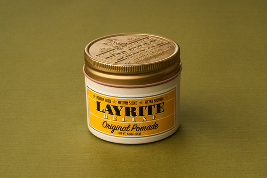 A jar of Layrite Original Pomade with a gold lid rests on a green surface. The yellow and white label features black and gold text, highlighting its medium hold, medium shine, and water-soluble formula—ideal for anyone seeking the classic experience of traditional pomade in a modern, water-based hair product.