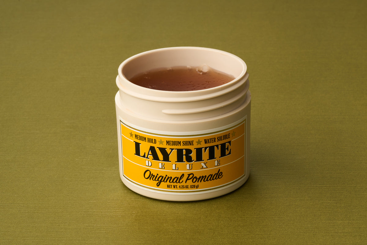 An open beige container of Layrite Original Pomade with a yellow label rests on a green surface, revealing the light brown gel inside—a great option for those looking for traditional, water-based hair styling products.