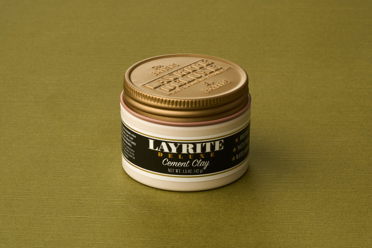 A small jar of Layrite Cement Hair Clay, a water-based pomade with a gold lid, sits on a green textured surface. The black label features white and gold text, emphasizing its reputation as an extreme hold hair product.