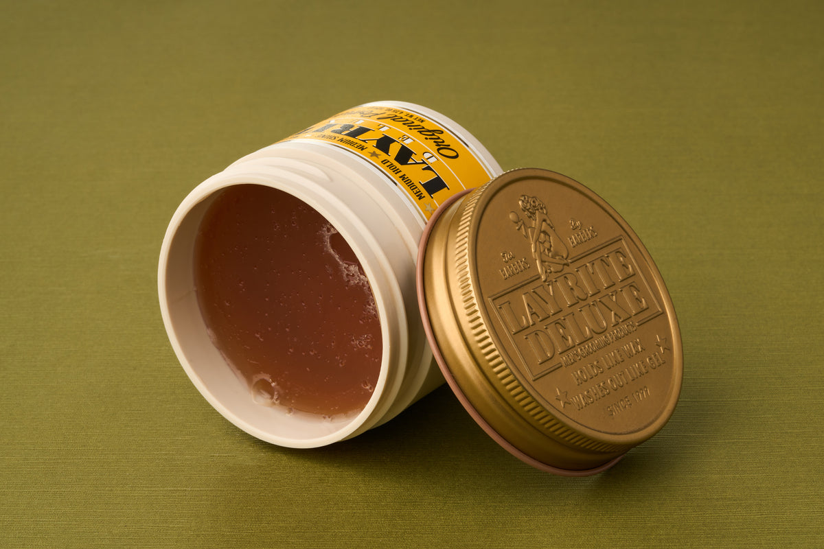 An open container of KIWI Leather Deluxe shoe polish with a brownish gel inside, lying on its side on a green surface, with the gold lid placed next to it—the texture and sheen reminiscent of Layrite Original Pomade.