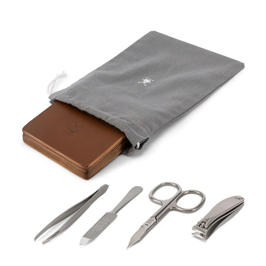 A gray drawstring pouch with a brown leather case partially inside is displayed above a row of stainless steel manicure tools—tweezers, nail file, cuticle pusher, and scissors—all neatly arranged as part of the Mühle 4pc Travel Manicure Set.