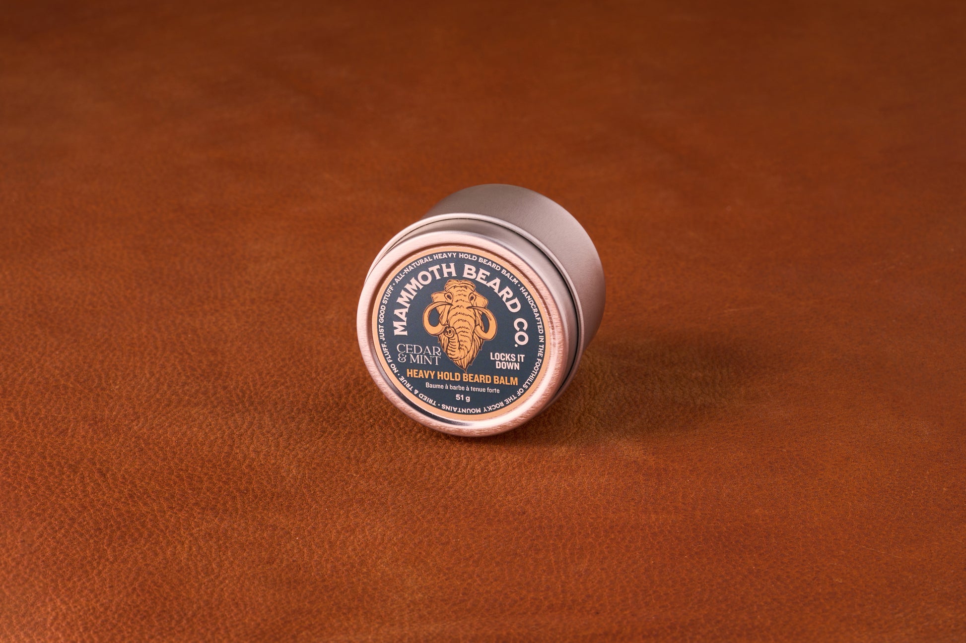 Mammoth Beard Co. Beard Balm Cedar and Mint 60ml tin on leather surface, featuring a mammoth logo and Locks It Down label, ideal for moisturizing and taming beards.