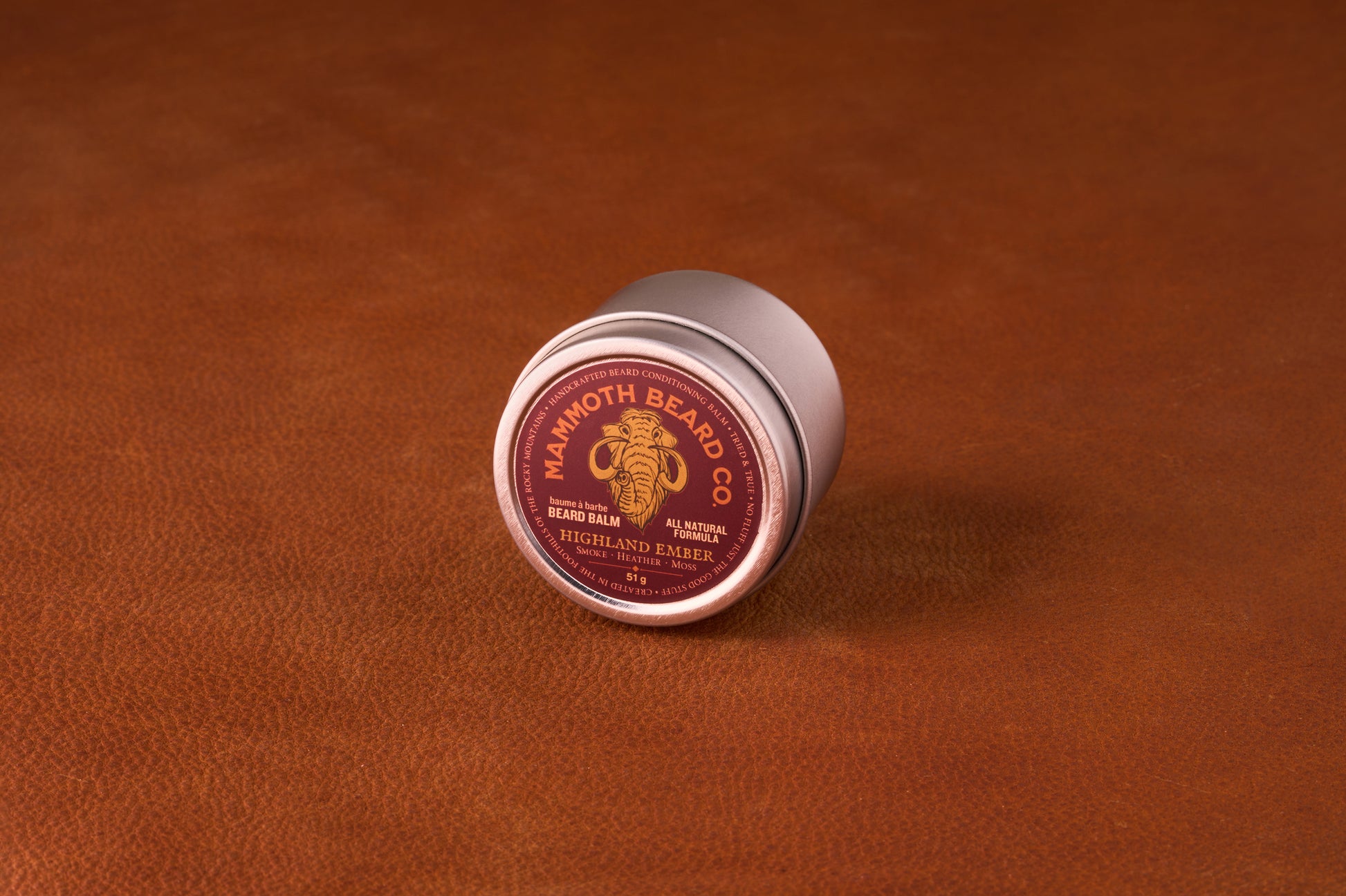 Mammoth Beard Co. Beard Balm Highland Ember tin on leather surface, offering natural beard care with argan and jojoba oils, shea butter, and beeswax for moisturizing and control.