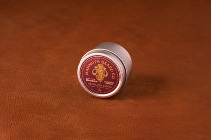 Mammoth Beard Co. Beard Balm Highland Ember tin on leather surface, offering natural beard care with argan and jojoba oils, shea butter, and beeswax for moisturizing and control.