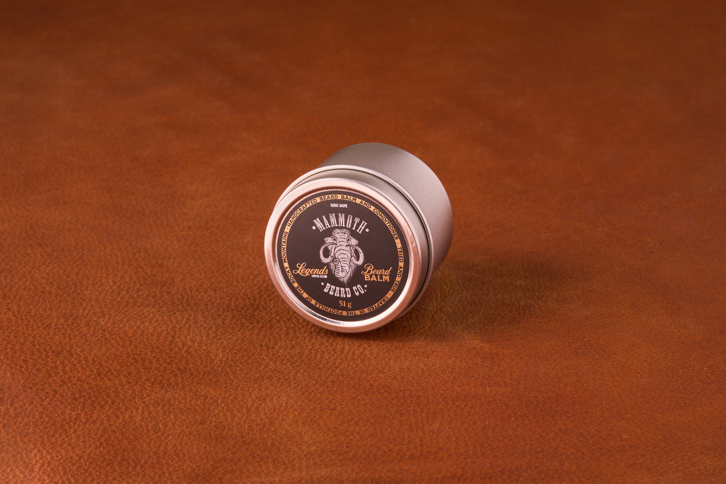 Mammoth Beard Co. Legends Beard Balm tin on leather, featuring a mammoth logo; moisturizes and controls beards with a unique Fougere scent blend.