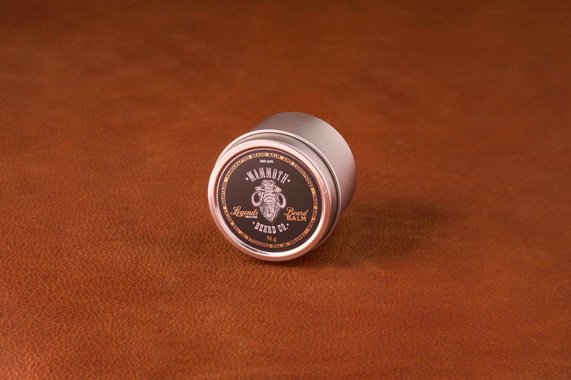 Mammoth Beard Co. Legends Beard Balm tin on leather, featuring a mammoth logo; moisturizes and controls beards with a unique Fougere scent blend.