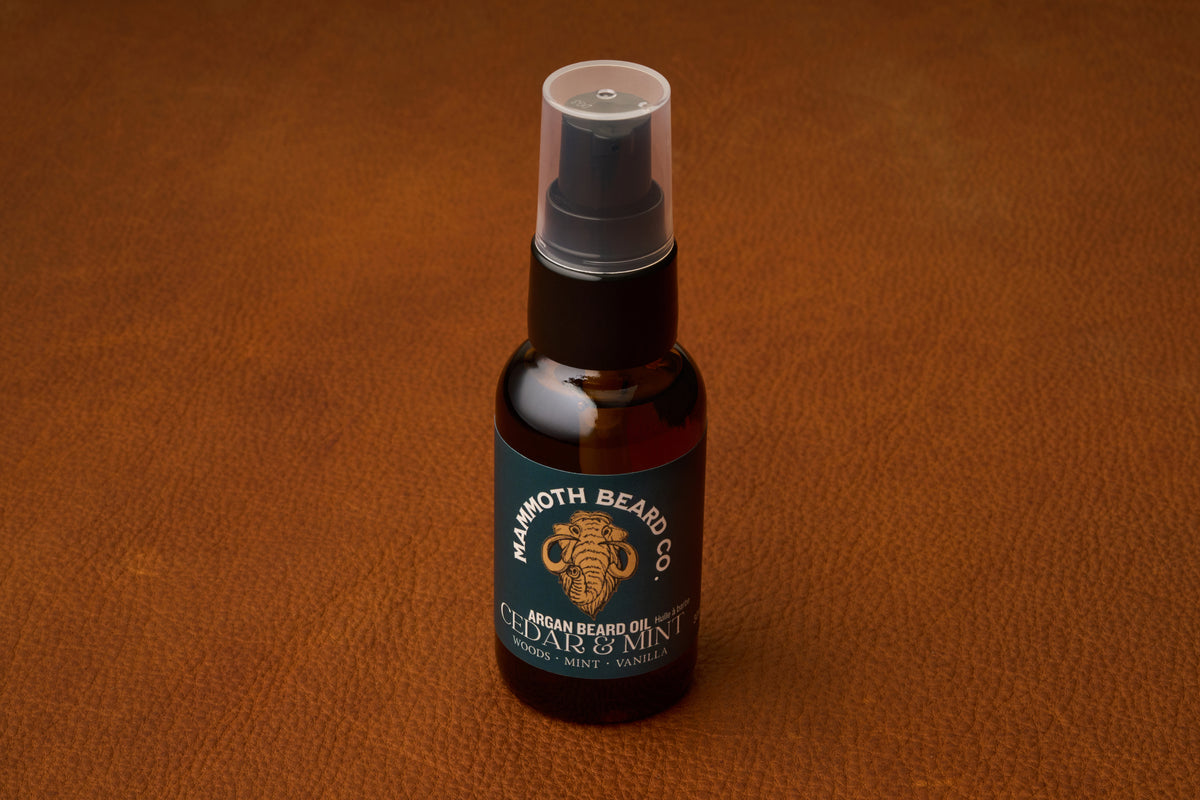 A 30ml amber bottle of Mammoth Beard Co. Beard Oil - Cedar and Mint, featuring a black label and pump, sits atop a brown leather surface—showcasing the brand’s commitment to premium, natural beard care.