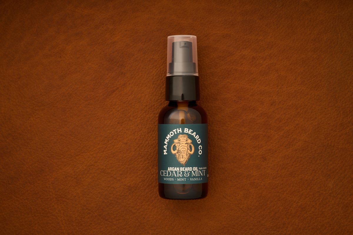 A bottle of Mammoth Beard Co. Beard Oil - Cedar and Mint 30ml, made with natural ingredients and featuring notes of woods, mint, and vanilla, rests on brown leather. The label showcases a mammoth illustration, highlighting the premium quality of this beard care product.
