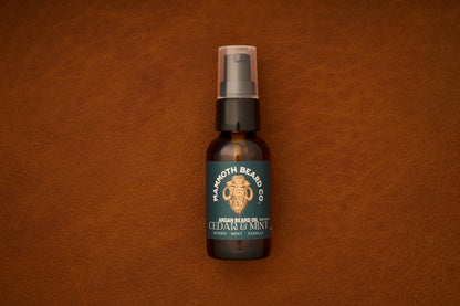 A bottle of Mammoth Beard Co. Beard Oil - Cedar and Mint 30ml, made with natural ingredients and featuring notes of woods, mint, and vanilla, rests on brown leather. The label showcases a mammoth illustration, highlighting the premium quality of this beard care product.
