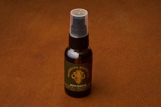 A bottle of Mammoth Beard Co. Beard Oil - Original Cedarwood 30ml with a spray cap rests on a textured brown leather surface, highlighting the premium quality of natural ingredients found in all their beard care products.