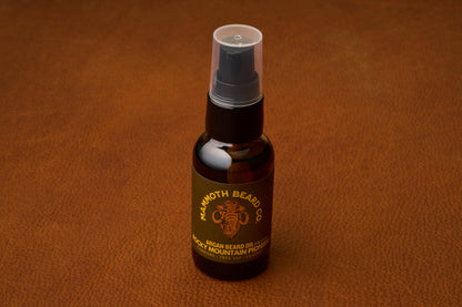 A small dark bottle of Mammoth Beard Co. Beard Oil - Rocky Mountain Pioneer 30ml with a pump lid rests on a brown textured surface. Crafted with natural ingredients, this premium beard care product features a gold mammoth logo and a label that reads Rocky Mountain Pioneer.