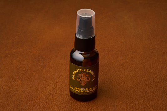 A small dark bottle of Mammoth Beard Co. Beard Oil - Rocky Mountain Pioneer 30ml with a pump lid rests on a brown textured surface. Crafted with natural ingredients, this premium beard care product features a gold mammoth logo and a label that reads Rocky Mountain Pioneer.