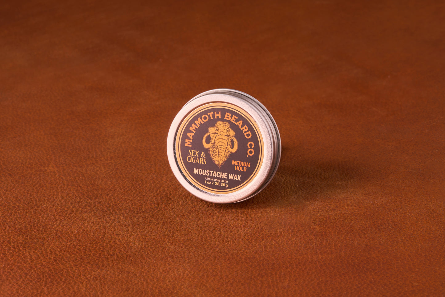 Mammoth Beard Co. Sex & Cigars Moustache Wax 1oz tin on leather surface, offering medium hold for versatile styling with a smoky, sweet scent.