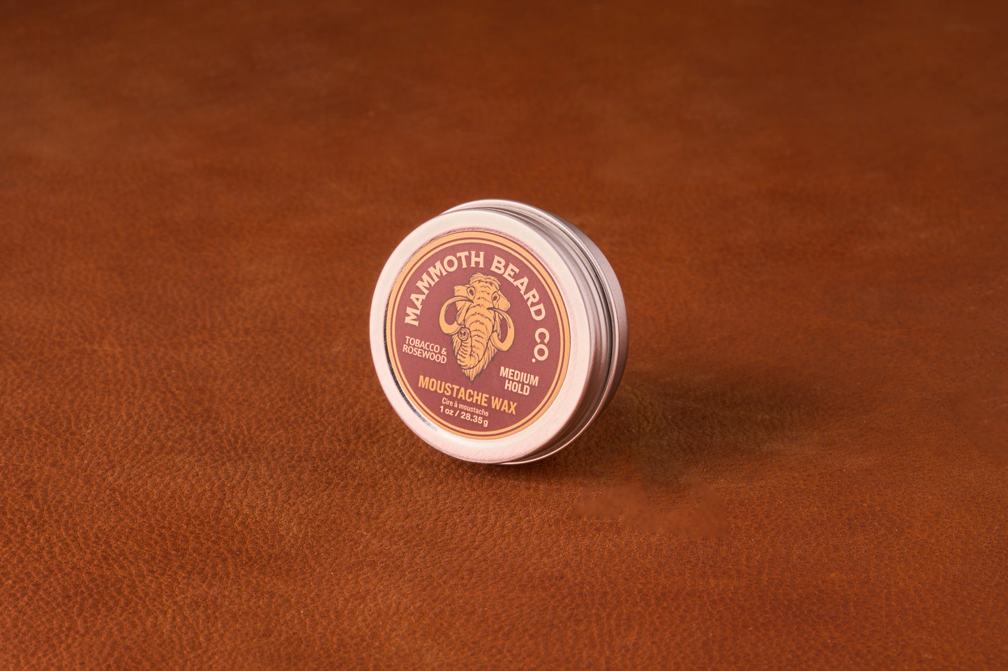 Mammoth Beard Co. Tobacco and Rosewood Moustache Wax 1oz tin on a leather surface; offers medium hold with a rich, slightly sweet scent.