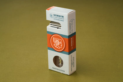 A Merkur Chrome DE Safety Razor, standing in its light gray packaging with orange accents and a helmeted profile logo, is displayed on a green surface. The box features the text “Merkur” and “Tradition & Design.”.