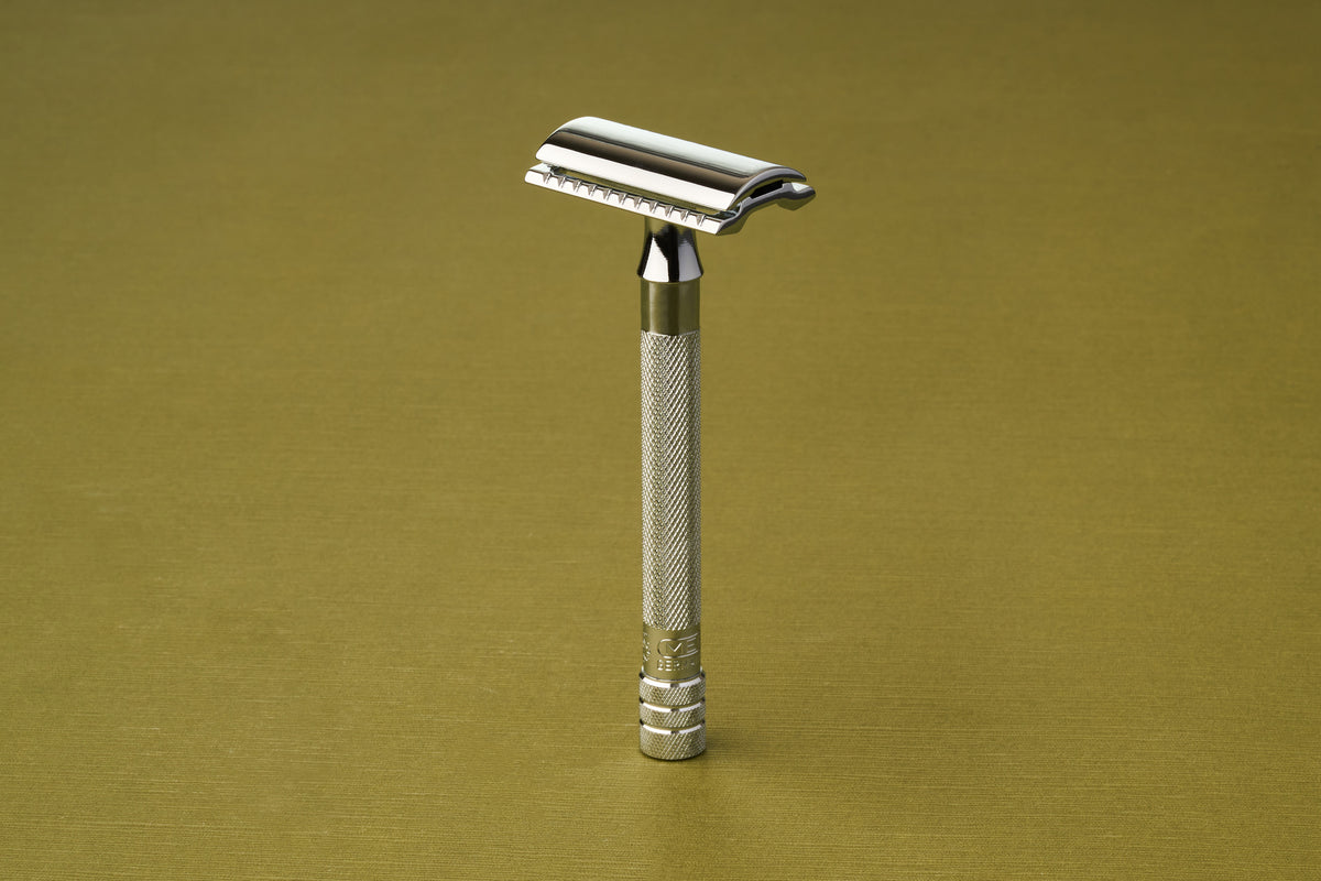 The Merkur Chrome DE Safety Razor, featuring a textured handle and chrome plating, stands upright against a light olive-green background.