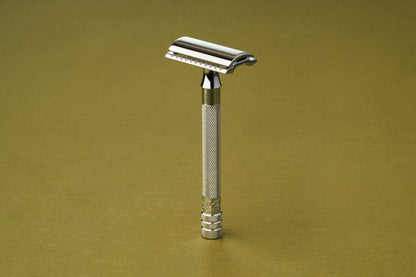 The Merkur Chrome DE Safety Razor, featuring a textured handle and chrome plating, stands upright against a light olive-green background.