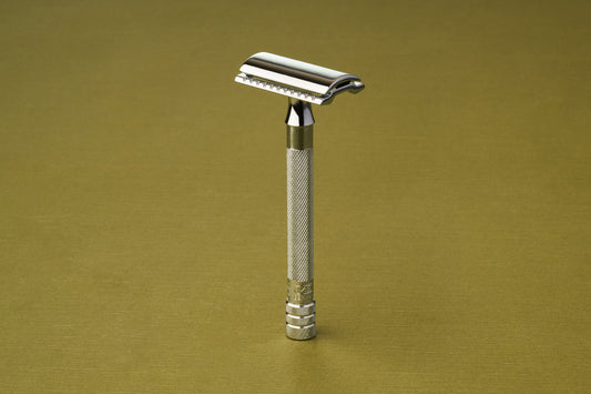 The Merkur Chrome DE Safety Razor, featuring a textured handle and chrome plating, stands upright against a light olive-green background.