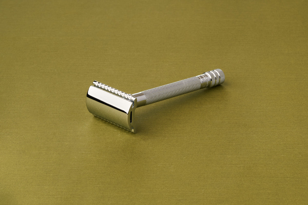 The Merkur Chrome DE Safety Razor, featuring a textured handle, rests at a slight angle on a smooth, gold-colored surface.