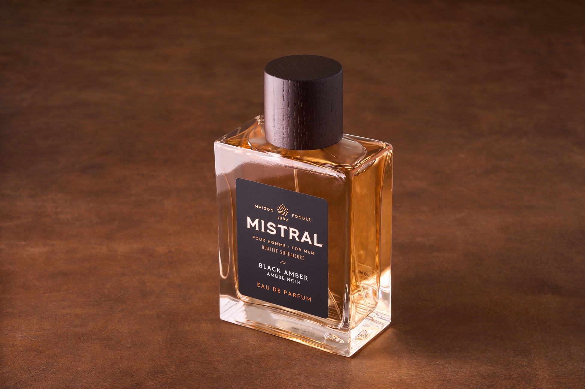 A 100ml bottle of Mistral Black Amber Eau de Parfum for men, featuring a dark wooden cap and black label with gold and white text, rests on a brown surface, showcasing its amber-colored liquid inside the glass bottle.