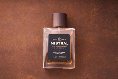 A bottle of Mistral Black Amber Eau de Parfum 100ml, a refined French men's fragrance with a dark wooden cap, rests on a brown textured surface. The label features the brand and Black Amber details in white and gold.
