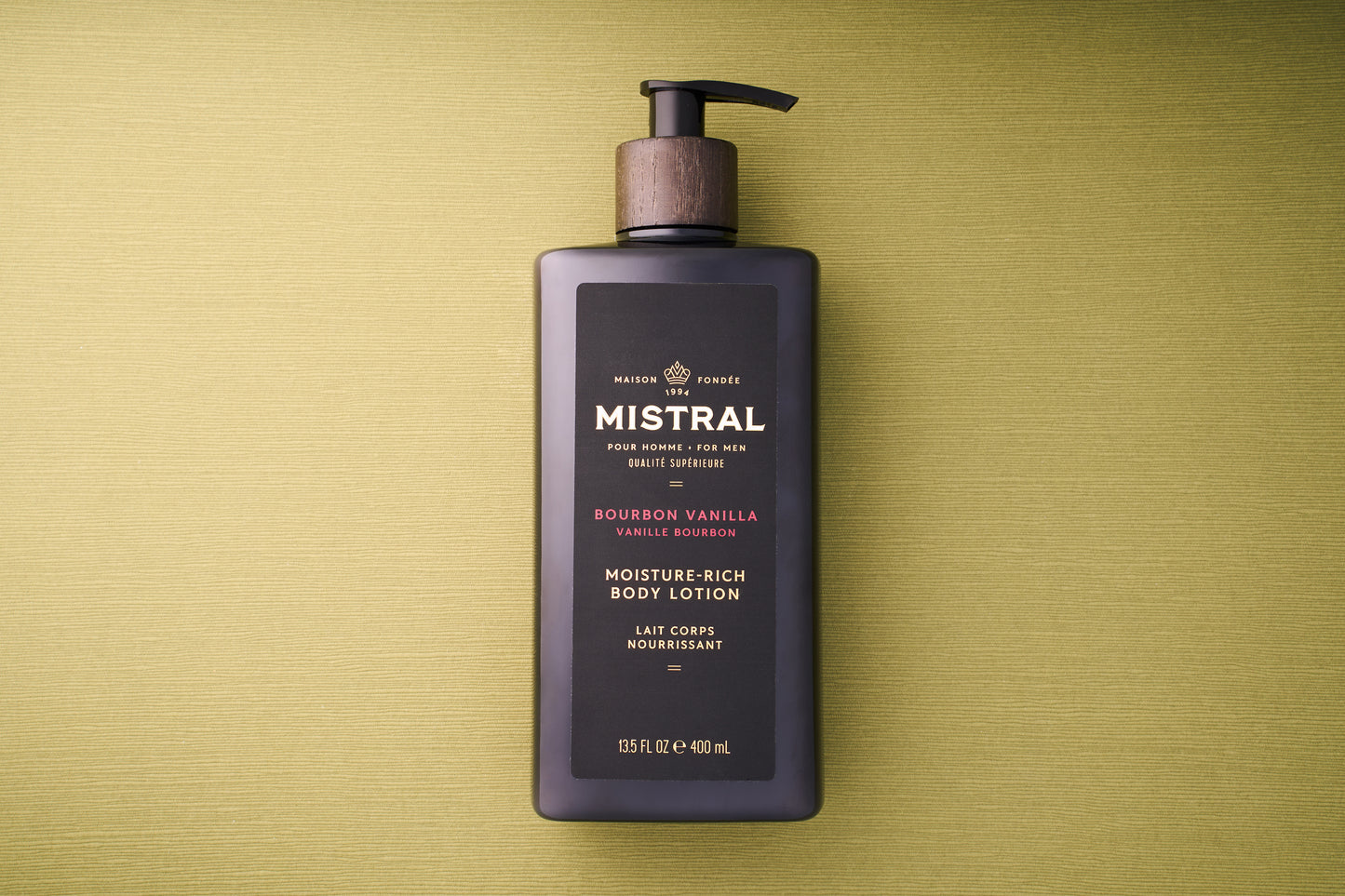 A black bottle of Mistral Body Lotion - Bourbon Vanilla with a wooden pump top sits on a textured green surface. The label features gold, white, and red accents, emphasizing the French fragrance for a touch of luxury.
