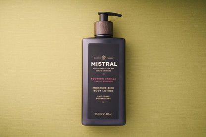 A black bottle of Mistral Body Lotion - Bourbon Vanilla with a wooden pump top sits on a textured green surface. The label features gold, white, and red accents, emphasizing the French fragrance for a touch of luxury.