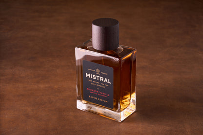 A bottle of Mistral Bourbon Vanilla Eau de Parfum 100ml rests on a brown surface, its amber fragrance glowing inside. The label features elegant gold and dark brown accents, topped with a dark wooden cap.