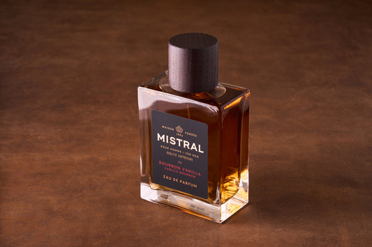 A bottle of Mistral Bourbon Vanilla Eau de Parfum 100ml rests on a brown surface, its amber fragrance glowing inside. The label features elegant gold and dark brown accents, topped with a dark wooden cap.