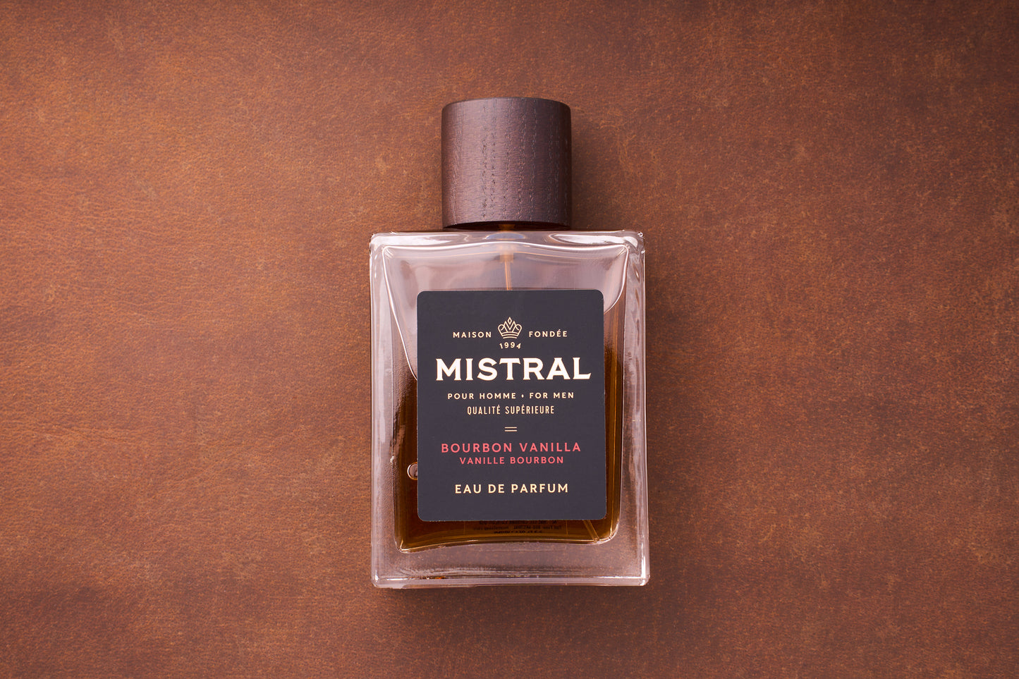 A 100ml bottle of Mistral Bourbon Vanilla Eau de Parfum for men, topped with a wooden cap, rests on a brown textured surface. Its black label displays gold and white text, highlighting this French-crafted fragrance.