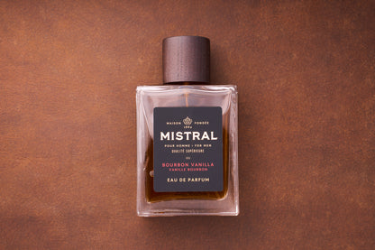A 100ml bottle of Mistral Bourbon Vanilla Eau de Parfum for men, topped with a wooden cap, rests on a brown textured surface. Its black label displays gold and white text, highlighting this French-crafted fragrance.