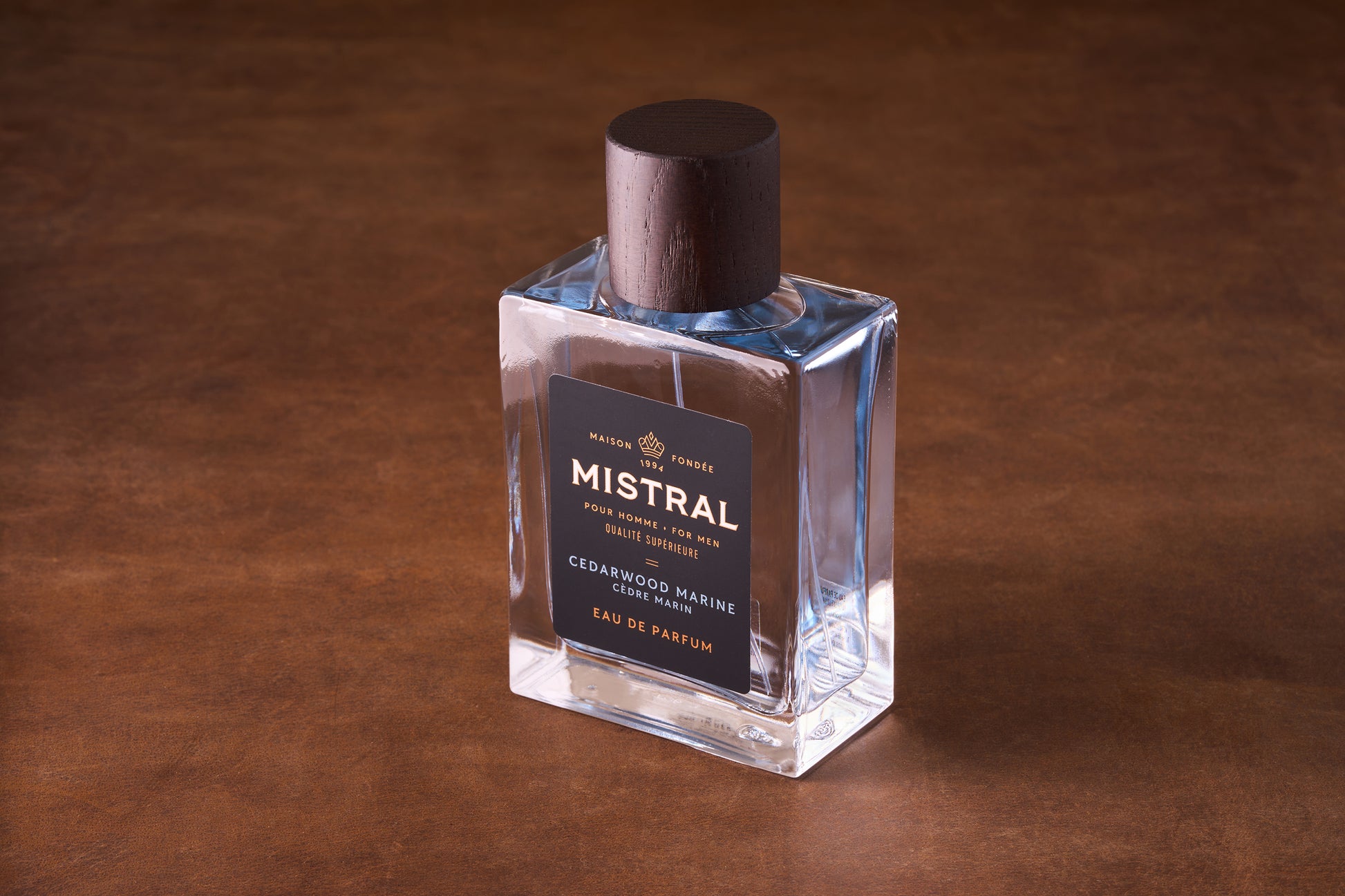 A clear glass bottle of Mistral Cedarwood Marine Eau de Parfum 100ml with a dark wooden cap rests on a brown surface. Its black label features gold and white text, reflecting its luxurious design.