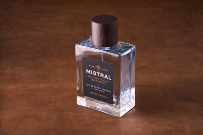 A clear glass bottle of Mistral Cedarwood Marine Eau de Parfum 100ml with a dark wooden cap rests on a brown surface. Its black label features gold and white text, reflecting its luxurious design.