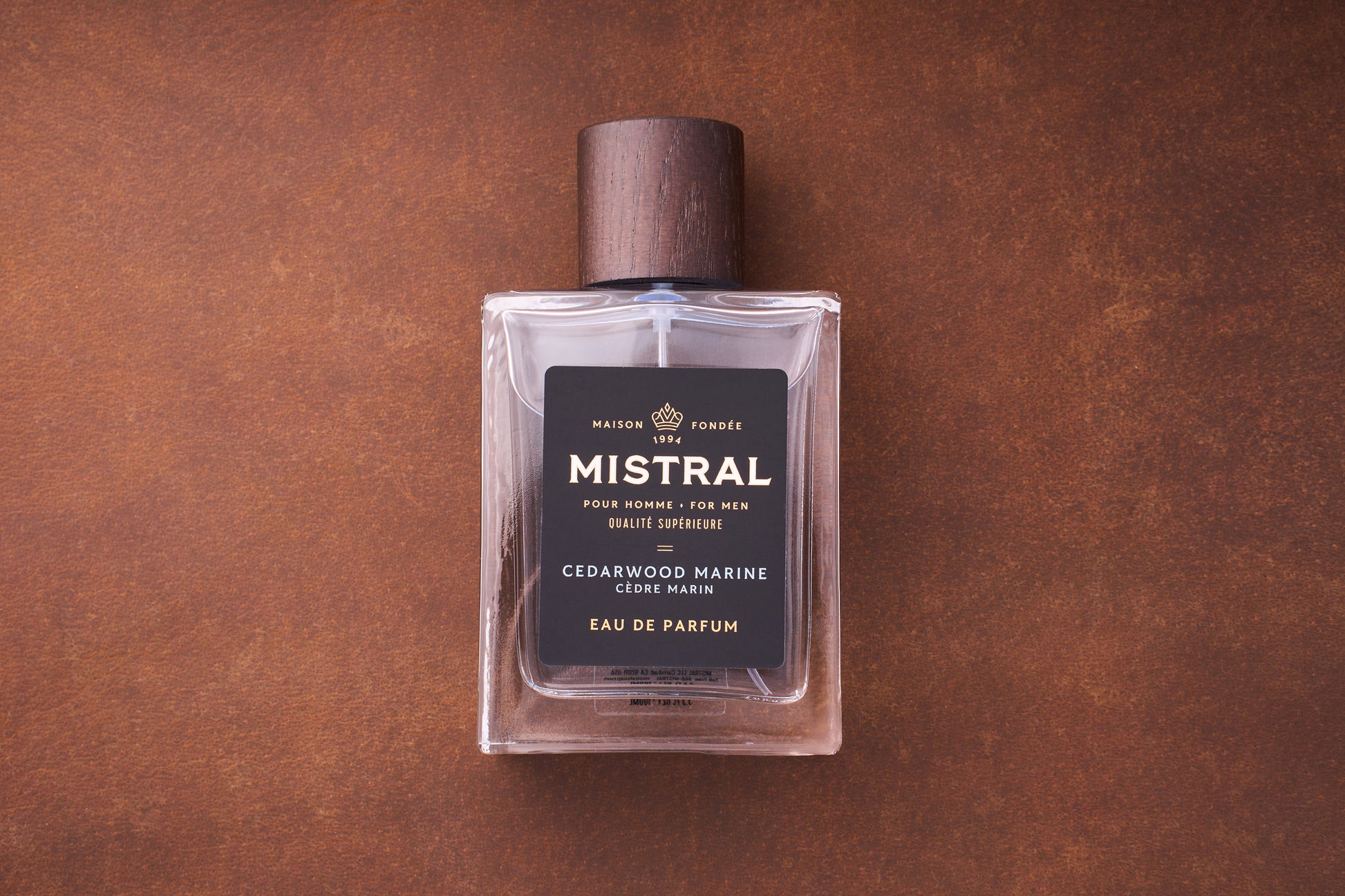 A 100ml bottle of Mistral Cedarwood Marine Eau de Parfum with a dark wooden cap and a black label featuring gold and white text sits on a brown textured surface, showcasing its French elegance.