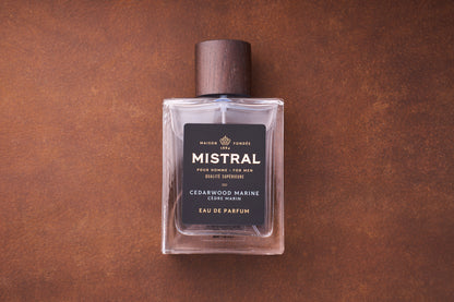 A 100ml bottle of Mistral Cedarwood Marine Eau de Parfum with a dark wooden cap and a black label featuring gold and white text sits on a brown textured surface, showcasing its French elegance.