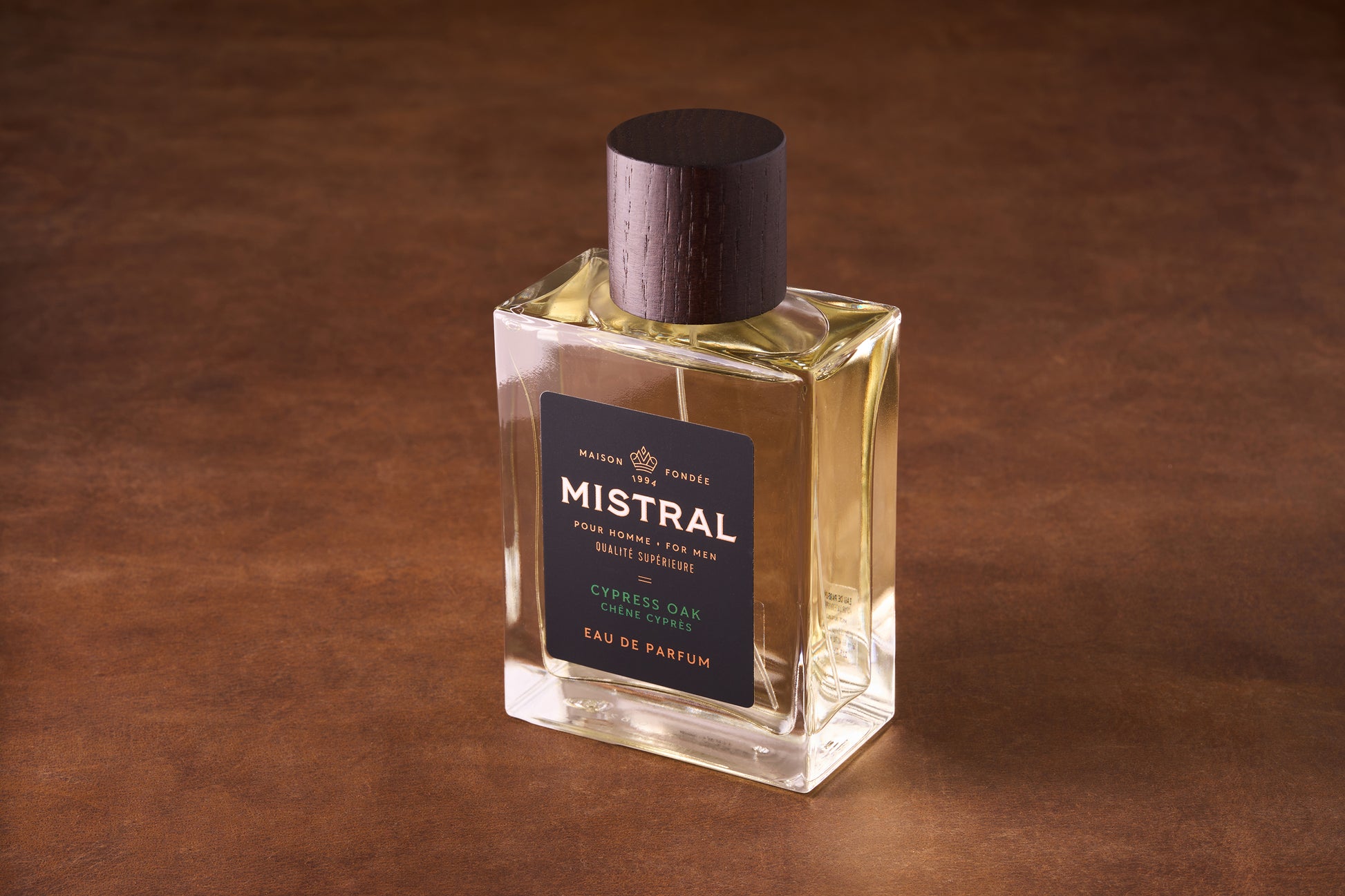A rectangular glass bottle of Mistral Cypress Oak Eau de Parfum 100ml with a dark wood cap sits on a brown surface. Its black label with gold and white text highlights this elegant French men's fragrance.