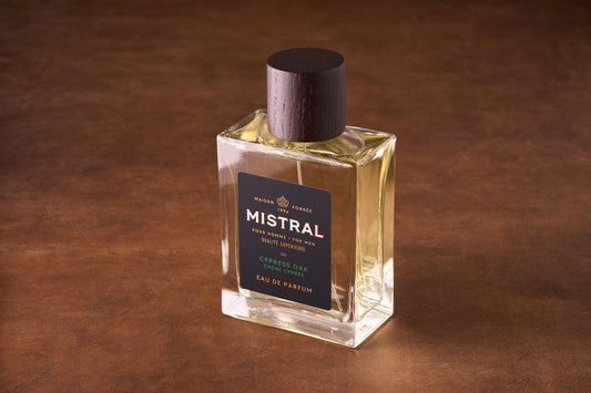 A rectangular glass bottle of Mistral Cypress Oak Eau de Parfum 100ml with a dark wood cap sits on a brown surface. Its black label with gold and white text highlights this elegant French men's fragrance.