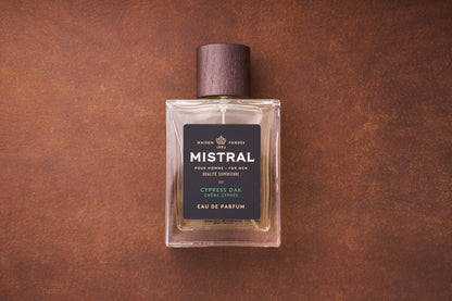 A 100ml bottle of Mistral Cypress Oak Eau de Parfum, a French men’s fragrance with a wooden cap and dark label, rests on a brown textured surface.