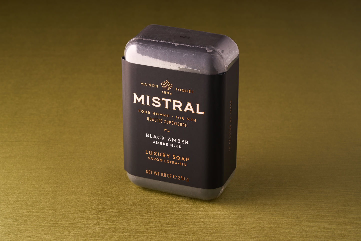 A rectangular Mistral For Men Black Amber Bar Soap, wrapped in black and gold packaging, rests on a plain olive green surface.
