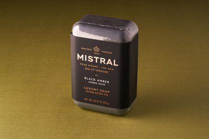 A rectangular Mistral For Men Black Amber Bar Soap, wrapped in black and gold packaging, rests on a plain olive green surface.