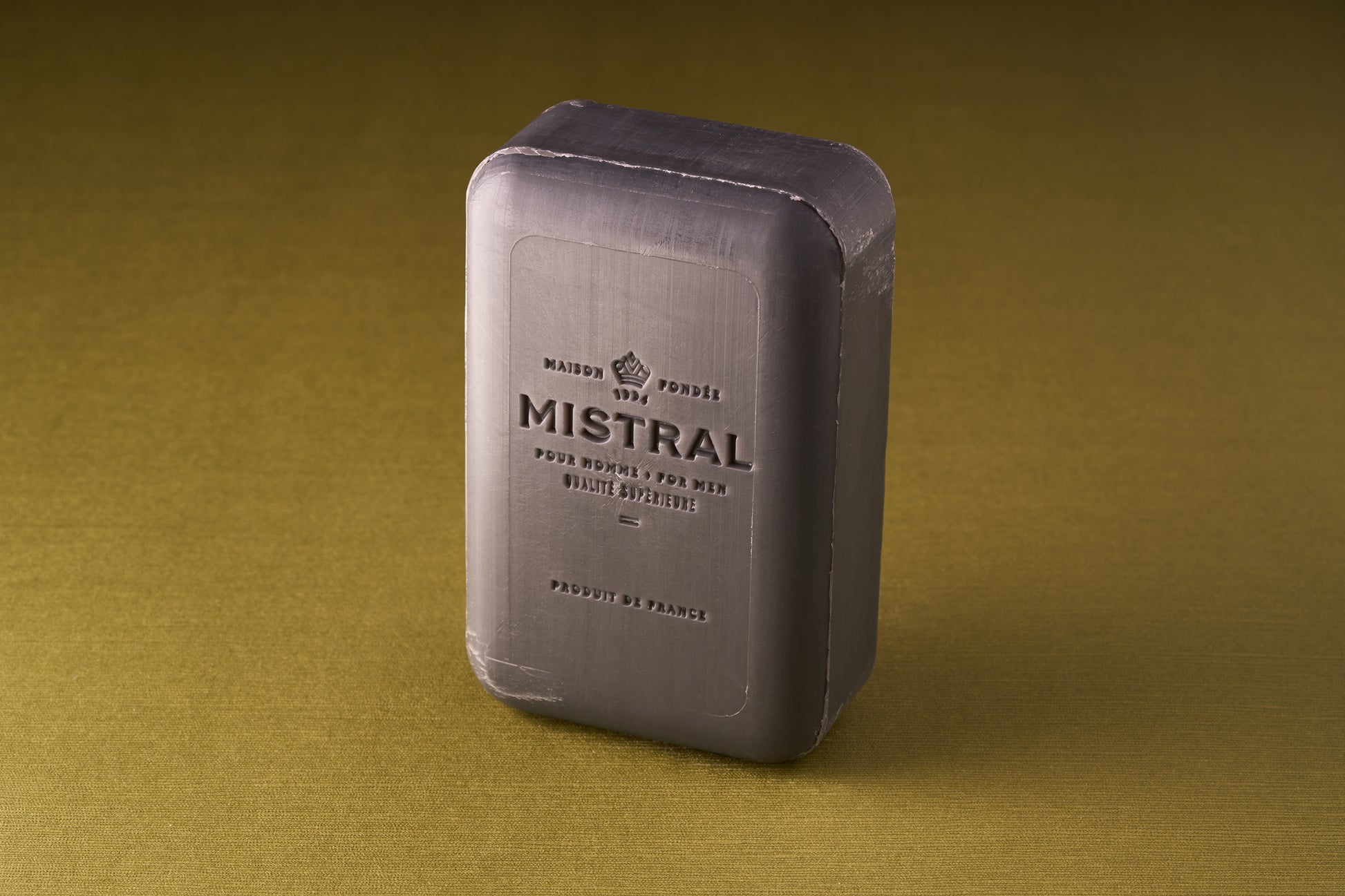 A rectangular bar of Mistral For Men Black Amber Bar Soap, triple-milled with French and Mistral text embossed on the front, stands on a textured olive-green surface.