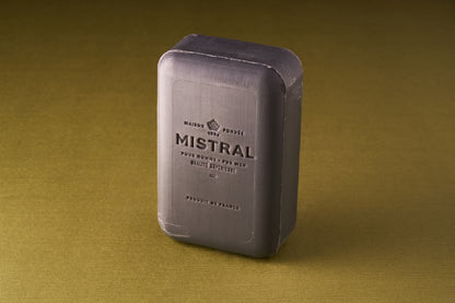 A rectangular bar of Mistral For Men Black Amber Bar Soap, triple-milled with French and Mistral text embossed on the front, stands on a textured olive-green surface.
