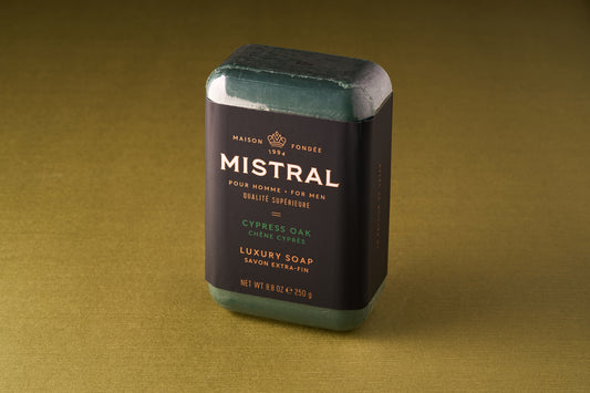 The Mistral For Men Cypress Oak Bar Soap is a rectangular green luxury soap labeled “Cypress Oak” with a black and gold wrapper, resting on a plain olive-green surface.