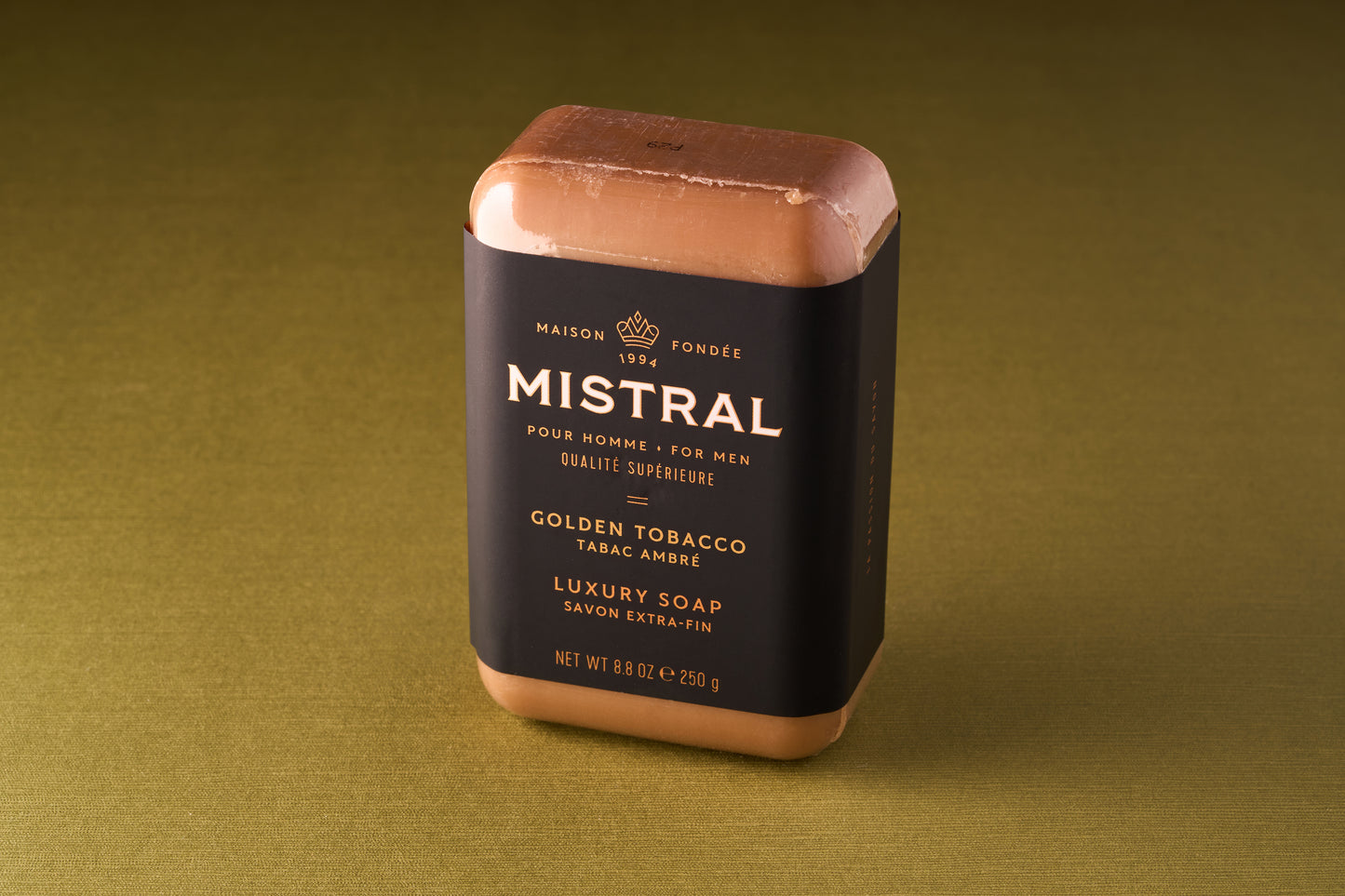 A Mistral For Men Golden Tobacco Bar Soap, triple-milled for lasting quality, is wrapped in black and gold packaging and stands upright on a greenish-brown surface.