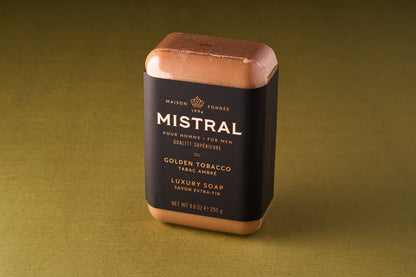 A Mistral For Men Golden Tobacco Bar Soap, triple-milled for lasting quality, is wrapped in black and gold packaging and stands upright on a greenish-brown surface.