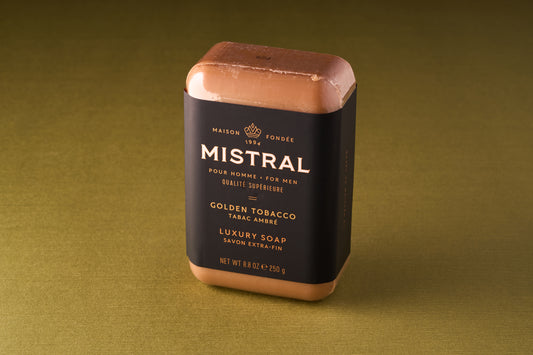 A Mistral For Men Golden Tobacco Bar Soap, triple-milled for lasting quality, is wrapped in black and gold packaging and stands upright on a greenish-brown surface.