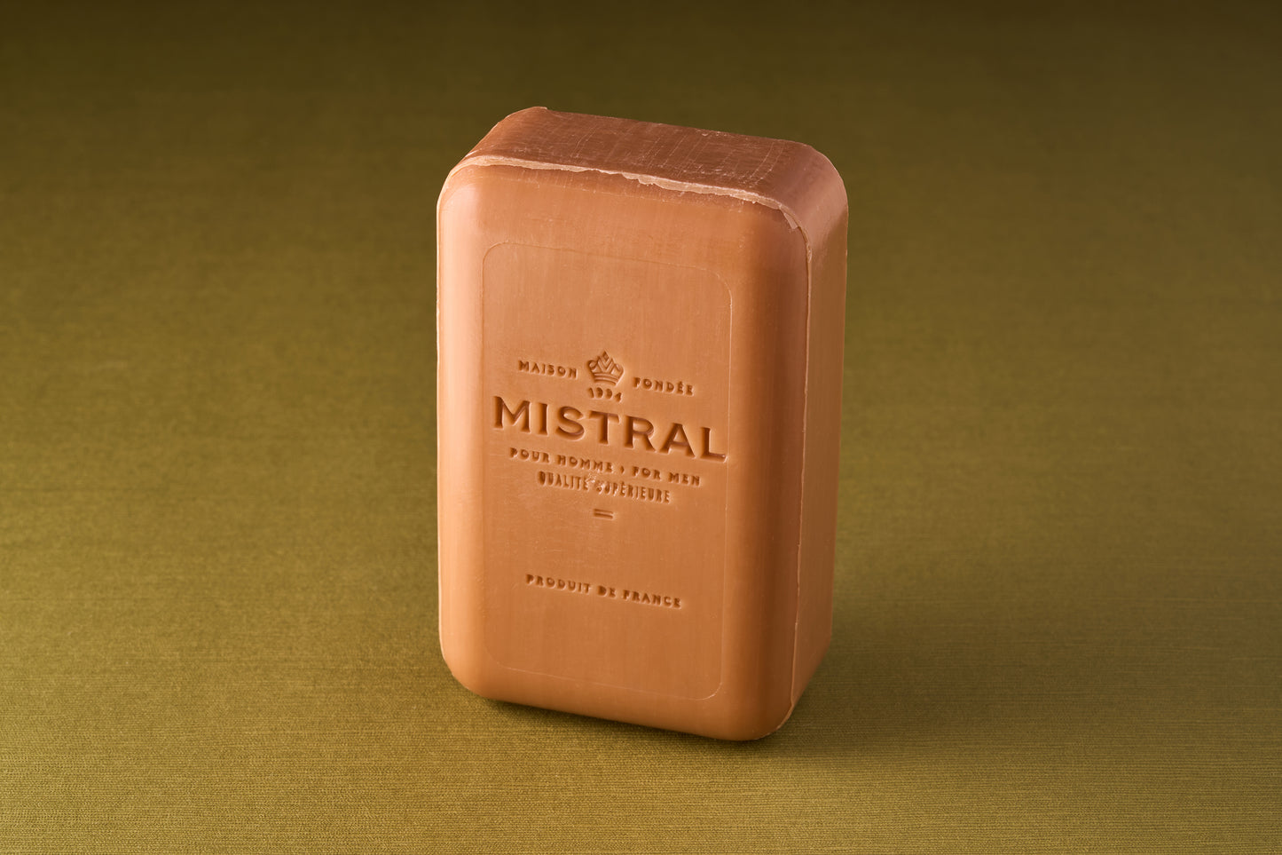 A rectangular Mistral For Men Golden Tobacco Bar Soap, light brown in color, stands upright on a textured olive green surface. The triple-milled soap is embossed with branding and French and English text.