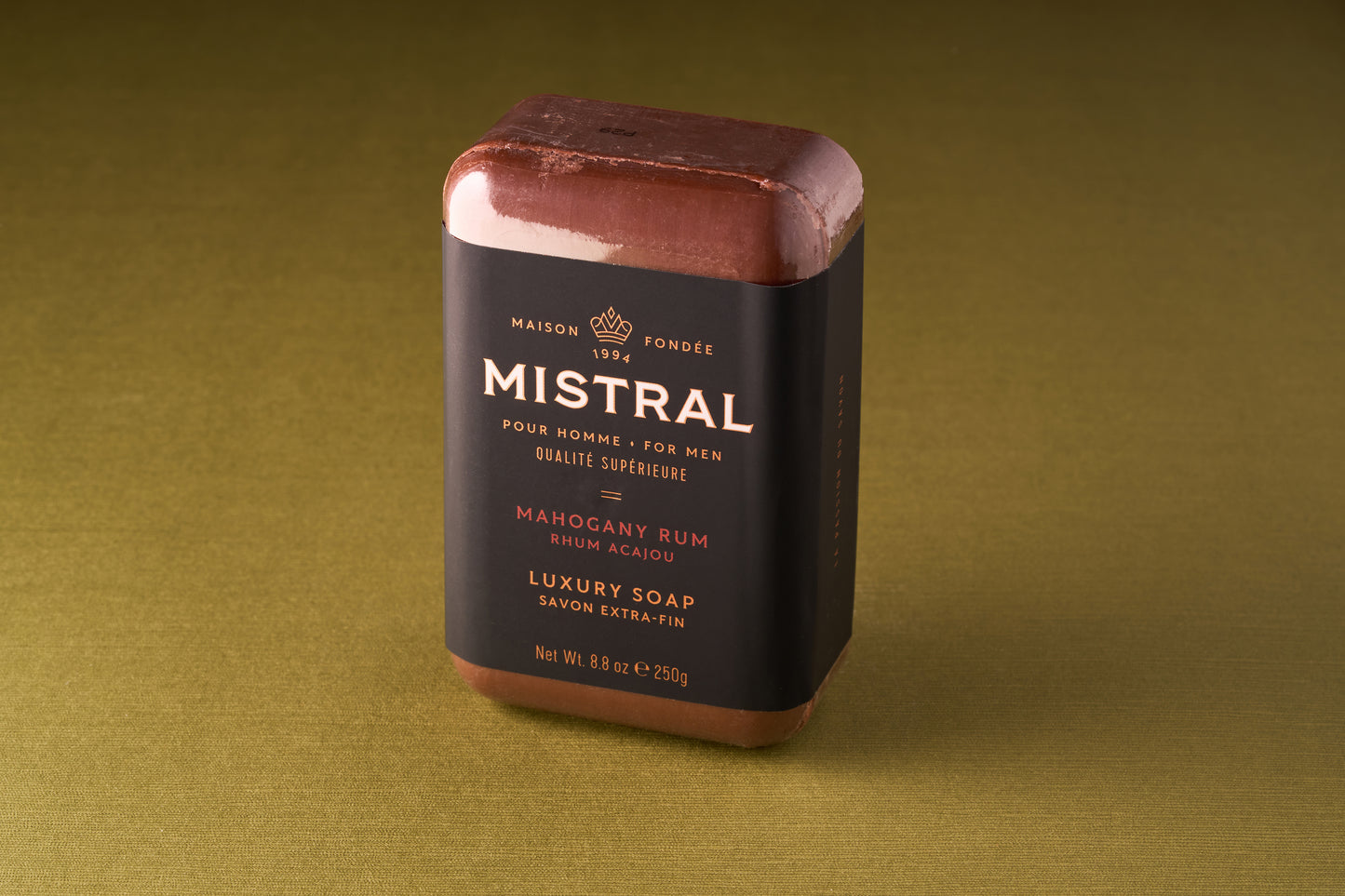 The Mistral For Men Mahogany Rum Bar Soap, a rectangular triple-milled bar with a dark wrapper featuring gold and white text, sits on an olive green surface, exuding a sophisticated mahogany rum scent.