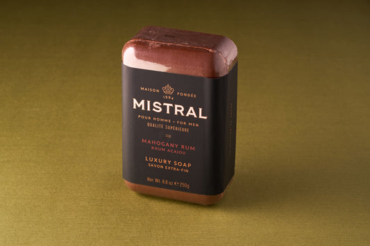 The Mistral For Men Mahogany Rum Bar Soap, a rectangular triple-milled bar with a dark wrapper featuring gold and white text, sits on an olive green surface, exuding a sophisticated mahogany rum scent.